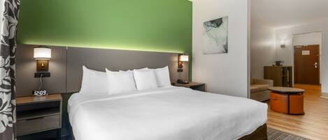 A hotel room with a large bed, two bedside tables with lamps, a clock, a green accent wall, a painting, a sofa, and a wooden floor.