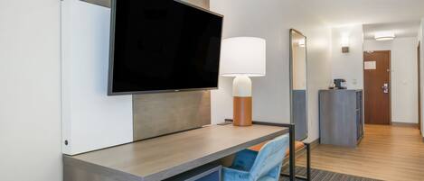 A hotel room with a flat-screen TV mounted on the wall, a desk with a chair, a lamp, a mirror, and a small cabinet.
