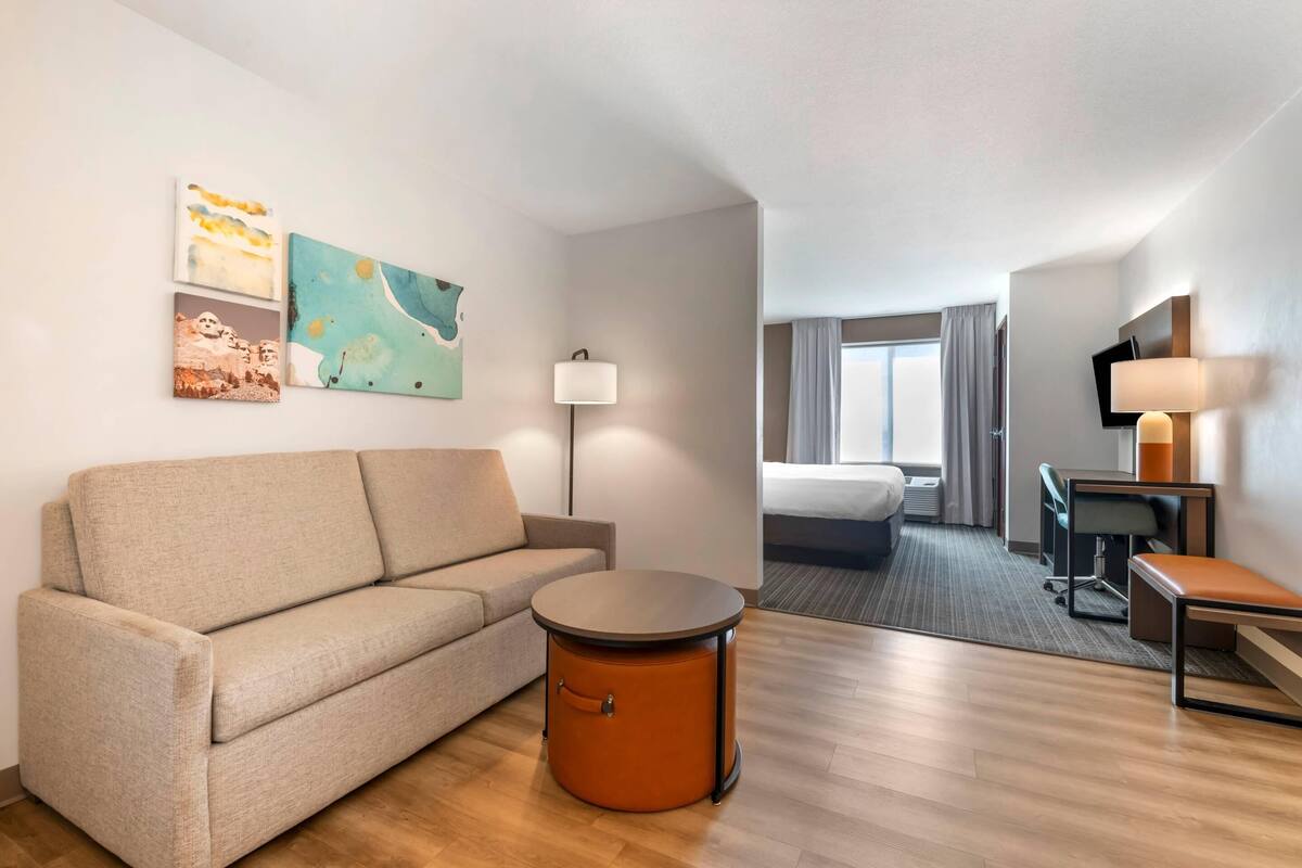 A hotel room with a sofa, a round table, a desk, a chair, a TV, and artwork on the wall.