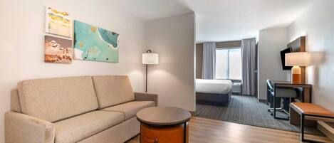 A hotel room with a sofa, a round table, a desk, a chair, a TV, and artwork on the wall.