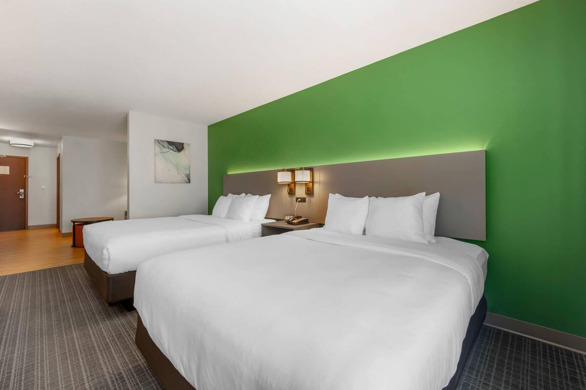 A hotel room with two beds, a green accent wall, a nightstand with a lamp, and a painting on the wall.