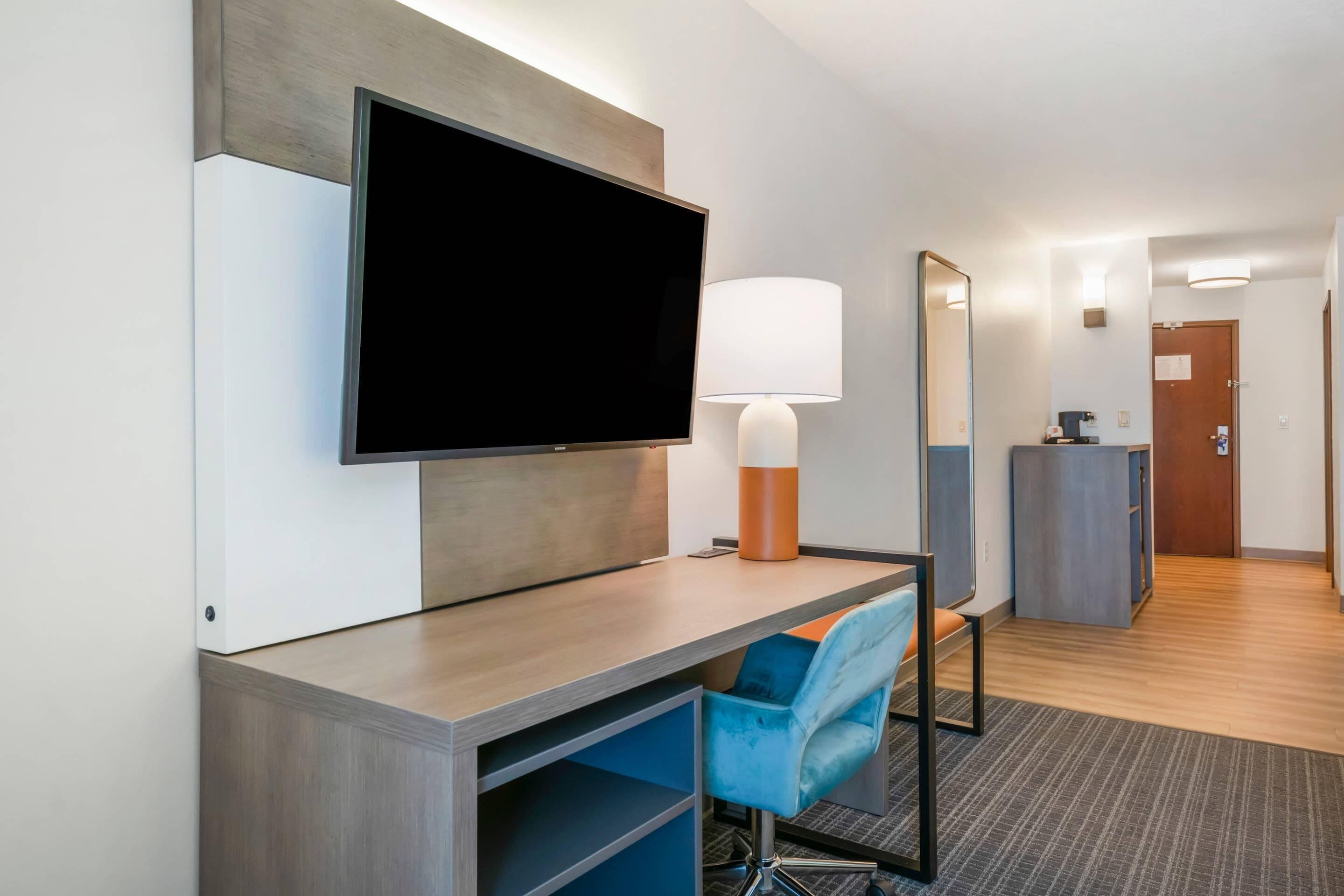 Habitación de hotel con televisor de pantalla plana, un escritorio con silla, lámpara, espejo y una pequeña kitchenette.