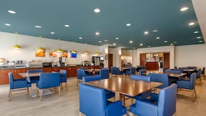 Ein geräumiger Essbereich mit mehreren Tischen und blauen Stühlen, ein Küchenbereich mit modernen Geräten sowie eine Decke mit Einbaustrahlern.