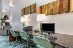 Habitación de hotel con escritorio, dos sillas verdes, computadora, lámpara y una obra de arte en la pared.