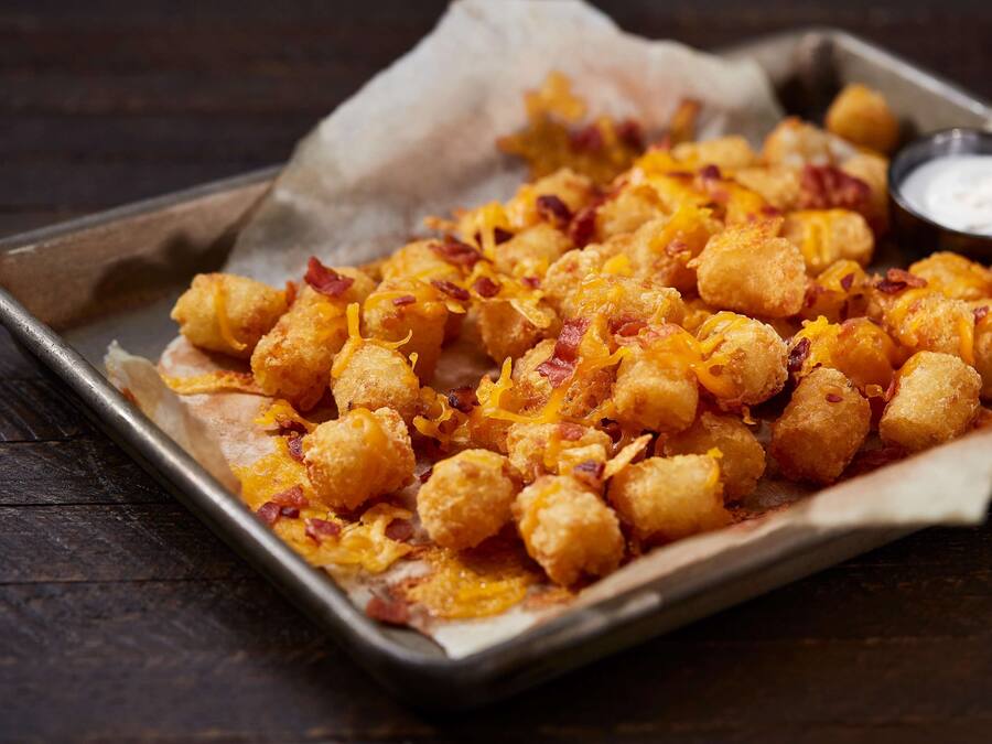 A tray of tater tots topped with cheese and bacon, served on a metal tray with a side of dipping sauce.