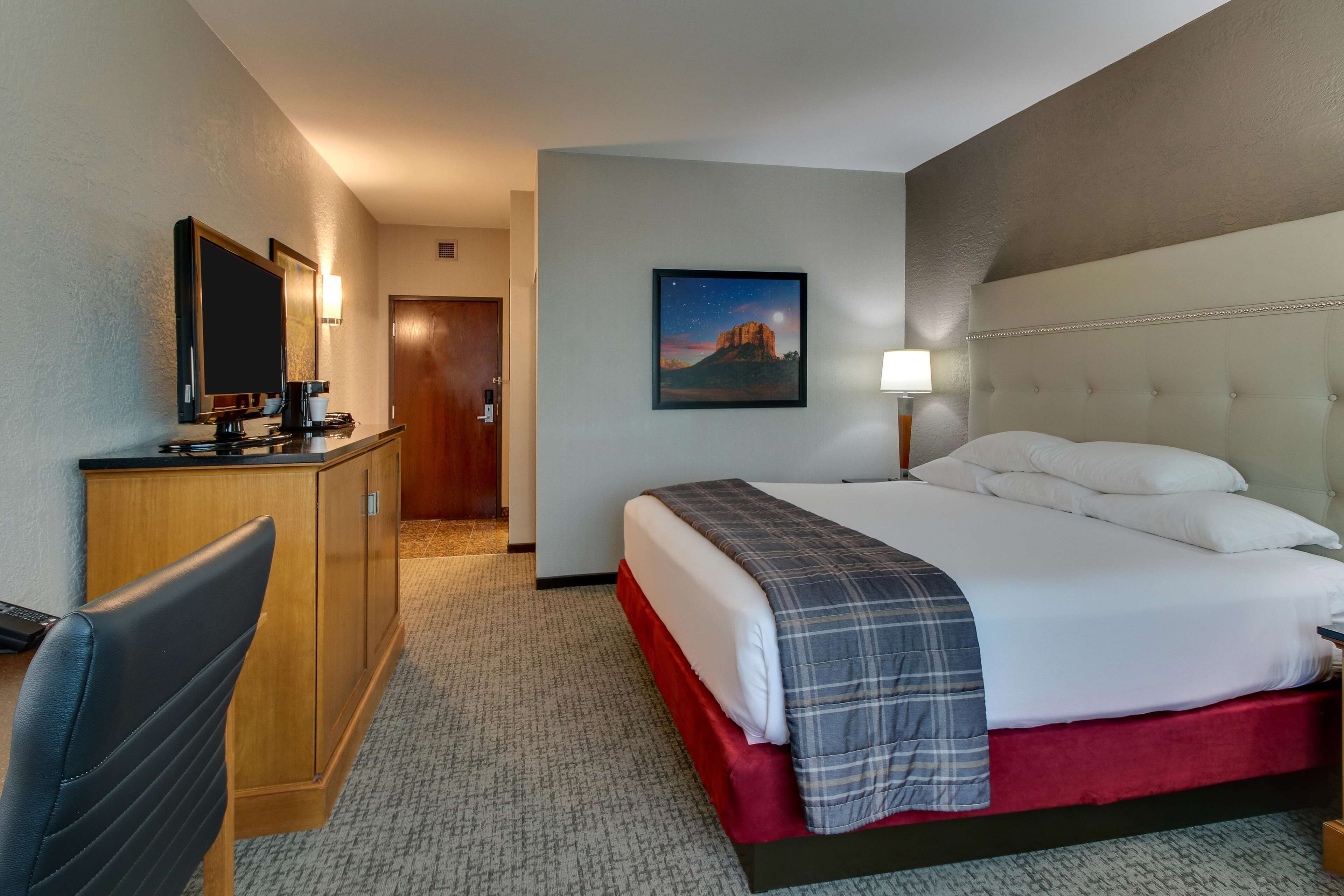 A hotel room with a bed, a desk, a chair, a painting, and a nightstand.
