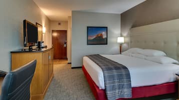 A hotel room with a bed, a desk, a chair, a painting, and a nightstand.