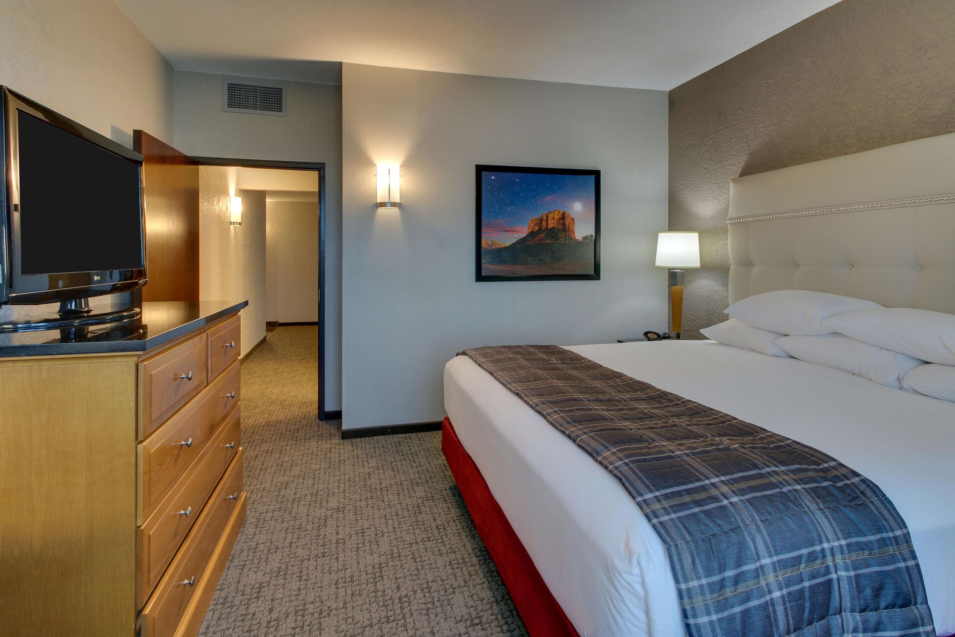 Habitación de hotel con cama, televisor sobre una cómoda de madera, mesita de noche con lámpara y un cuadro en la pared.