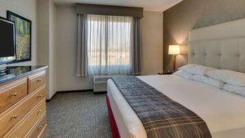 A hotel room with a bed, a television, a dresser, a window with curtains, and a lamp.