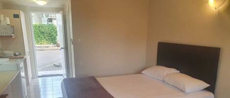 A hotel room with a bed, a bedside table, and a desk with a chair. There is a door leading to the outside.