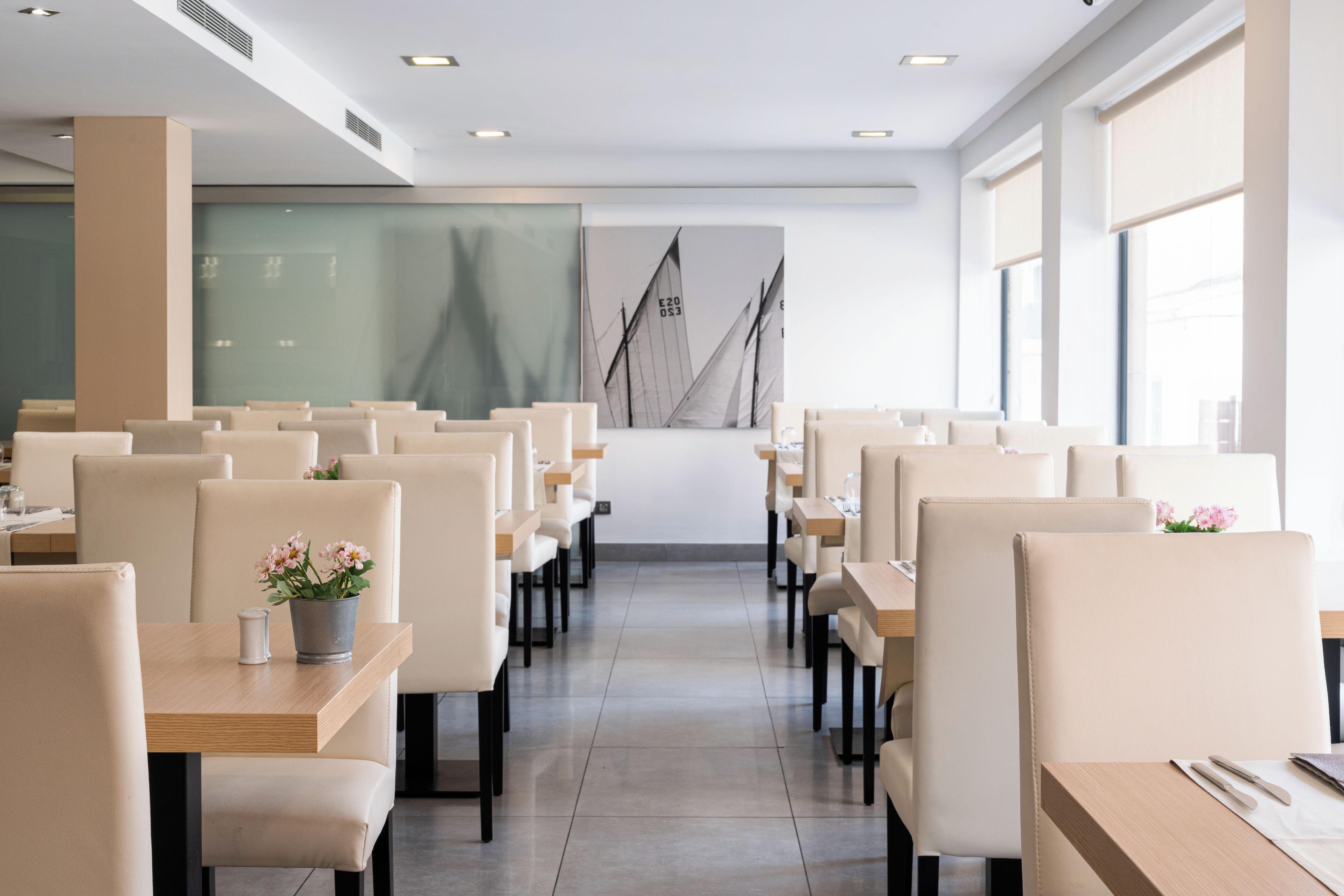 A modern dining area with rows of tables and chairs, a large window on the right, and a sailboat painting on the wall.
