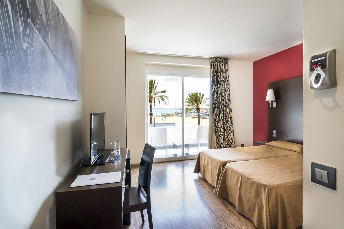 Een hotelkamer met een bureau, stoel, twee bedden, een televisie en een groot raam met uitzicht op palmbomen en een strand.