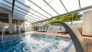 A swimming pool with a waterfall feature, surrounded by lounge chairs and a covered area with a glass roof. - Nautic Hotel & Spa (Palma de Mallorca)