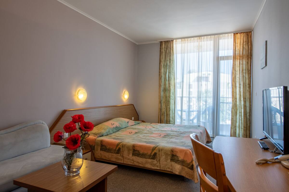 Une chambre d’hôtel avec un lit, un bureau en bois, une télévision et un vase rempli de fleurs rouges.