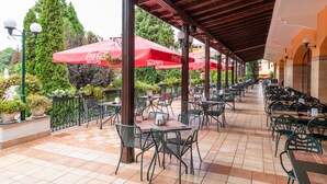 Ein Straßencafé mit roten Sonnenschirmen, Metallstühlen und -tischen, umgeben von Grünpflanzen und einem gefliesten Boden.