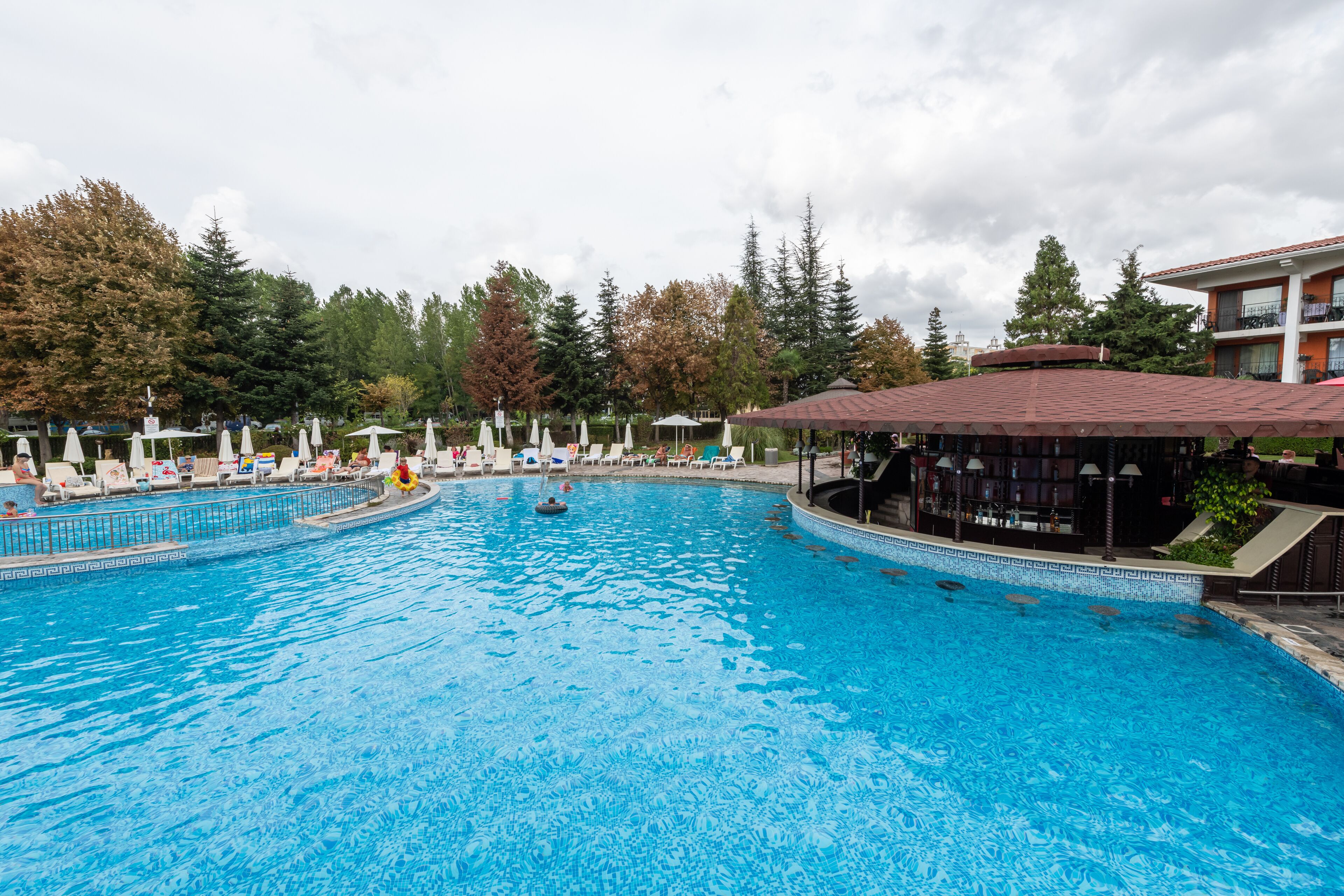 A large outdoor swimming pool with lounge chairs and umbrellas, surrounded by trees and a building with a red roof.