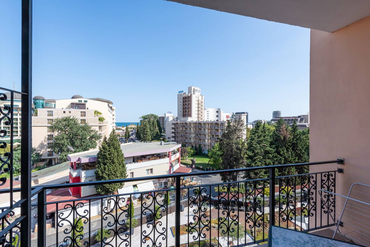 Un balcon doté de balustrades en fer forgé, offrant une vue sur un paysage urbain avec des bâtiments, des arbres et un ciel dégagé.