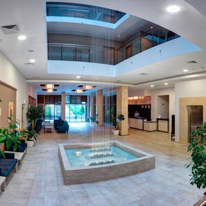 A modern hotel lobby with a central water feature, a reception desk, seating areas, and a glass ceiling. - Slavey (Golden Sands)