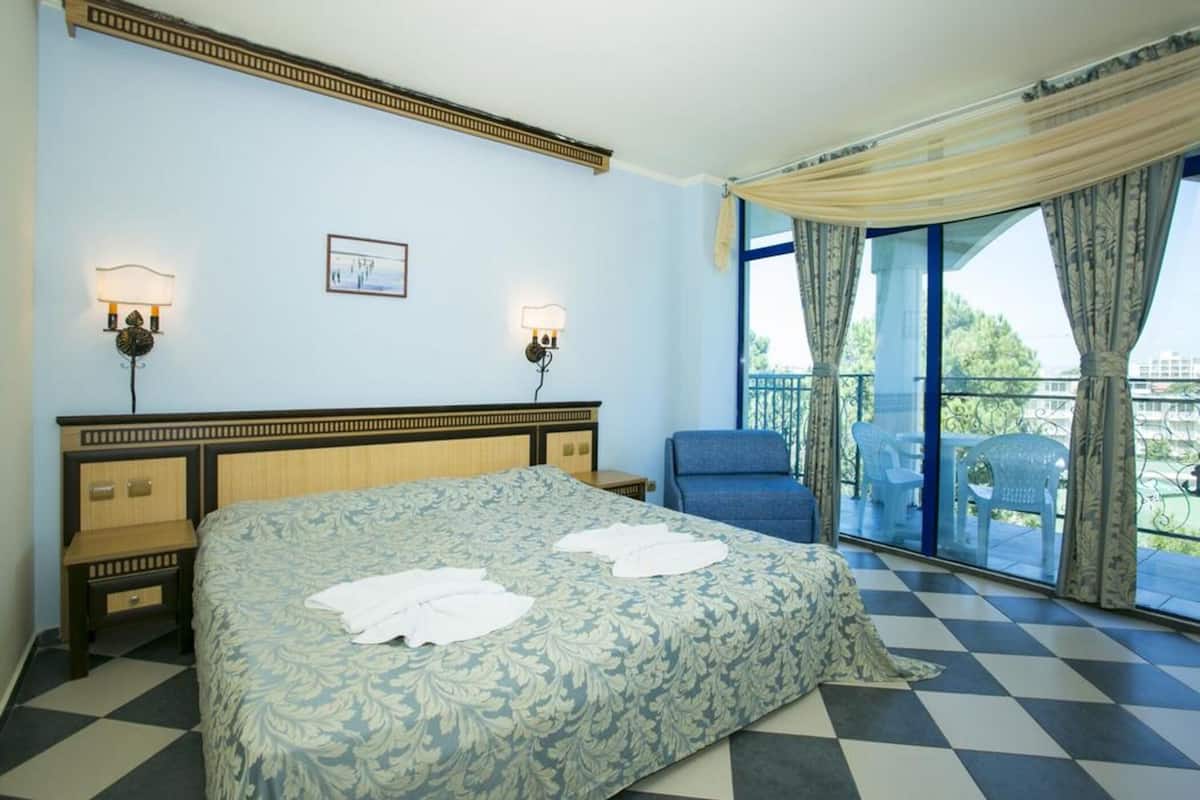 Una habitación de hotel con una cama, dos lámparas de noche, una silla, un balcón con cortinas y un suelo a cuadros.