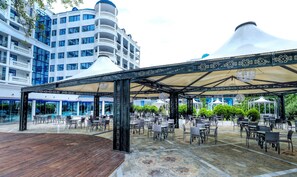 Uma área externa com mesas e cadeiras, um pavilhão coberto e uma piscina ao fundo.