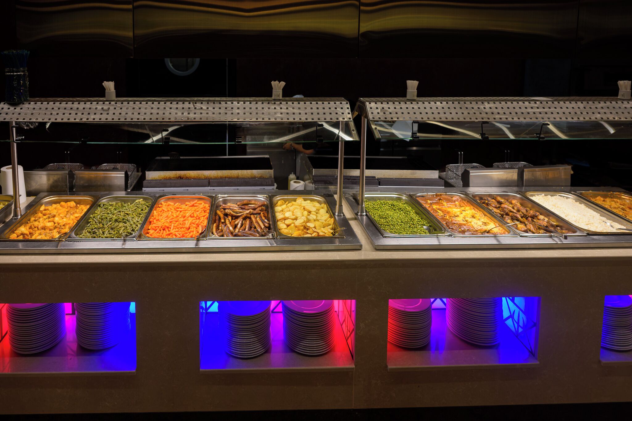 A buffet with various food options and a stack of plates underneath.