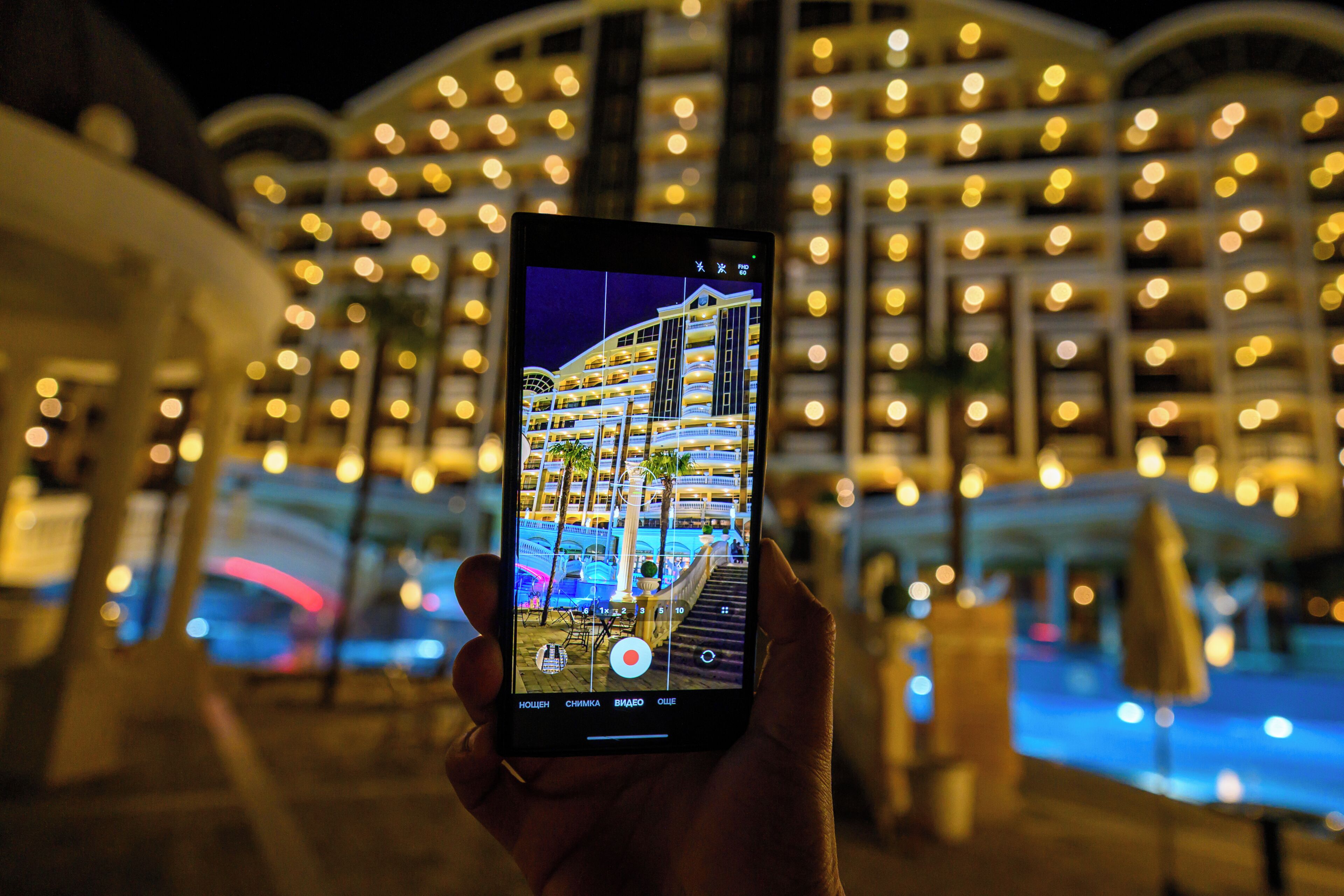 Eine Hand hält ein Smartphone, auf dem ein Hotel mit mehreren Balkonen und einem Poolbereich zu sehen ist.