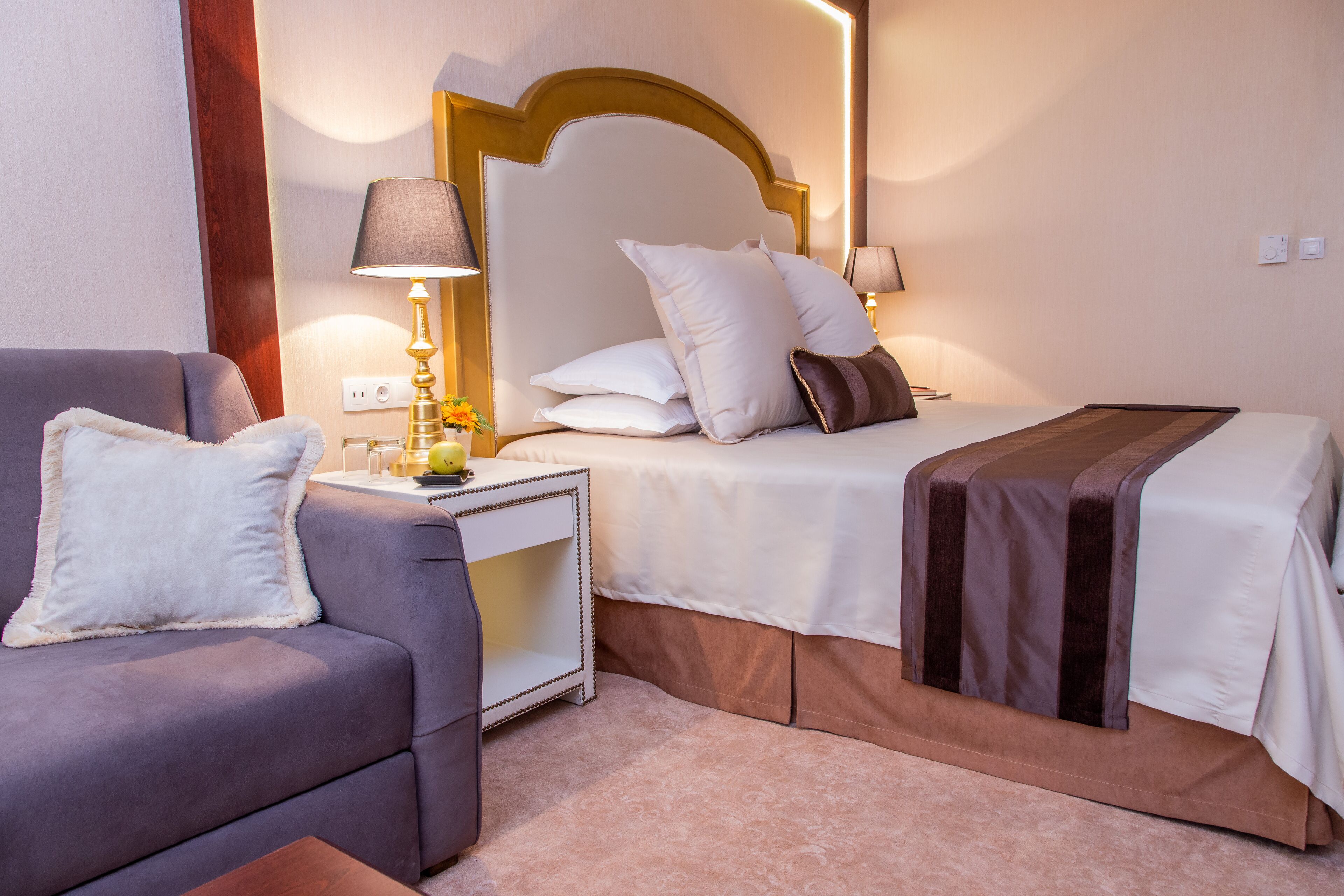 A hotel room with a bed, a sofa, a nightstand with a lamp, and a bedside table with a lamp.