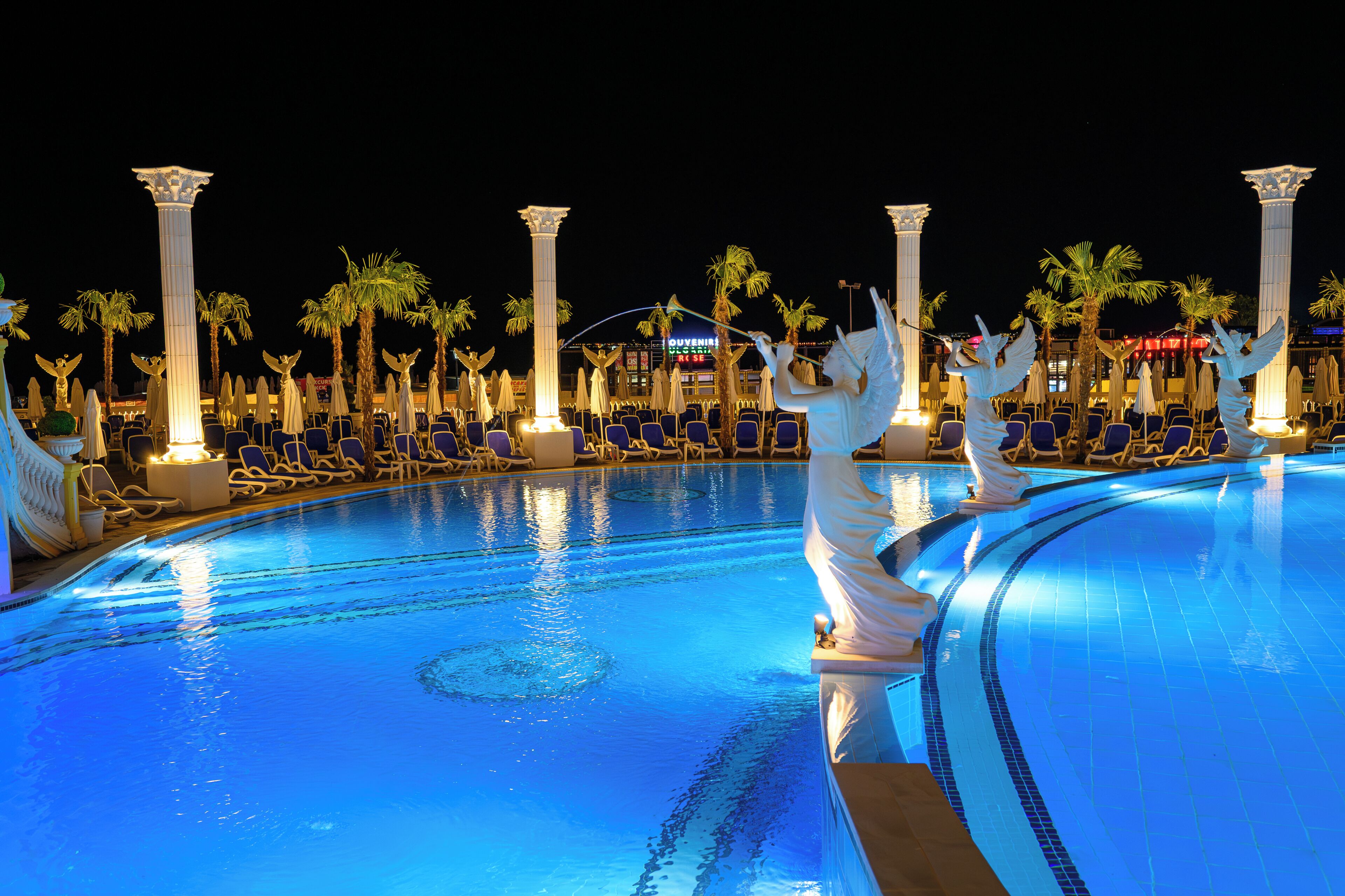 Una piscina con estatuas de ángeles, palmeras y sillas de descanso.