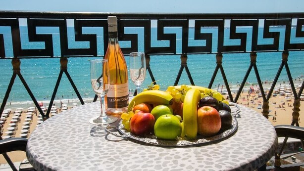 Une table avec une bouteille de vin, des verres et une assiette de fruits sur un balcon donnant sur une plage.