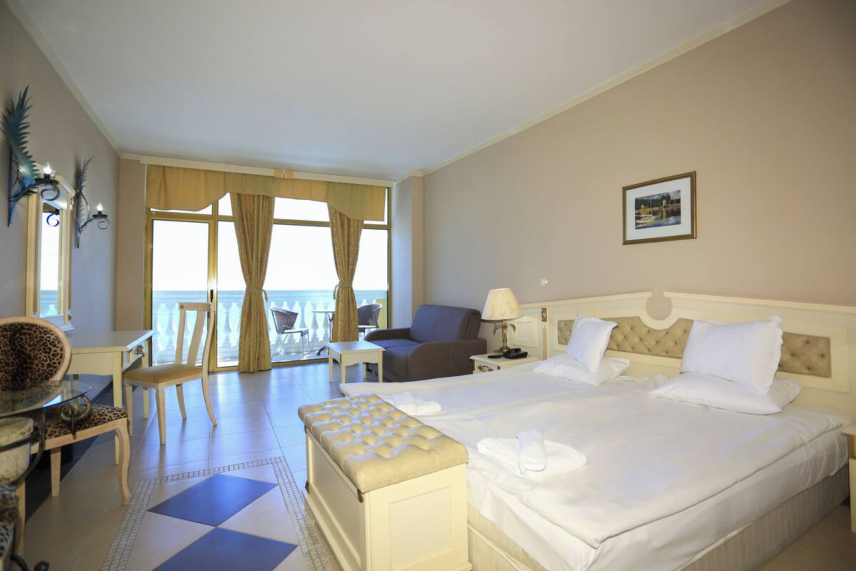 A hotel room with a large bed, a sitting area with a chair and a table, a view of the sea through a large window, and a framed picture on the wall.