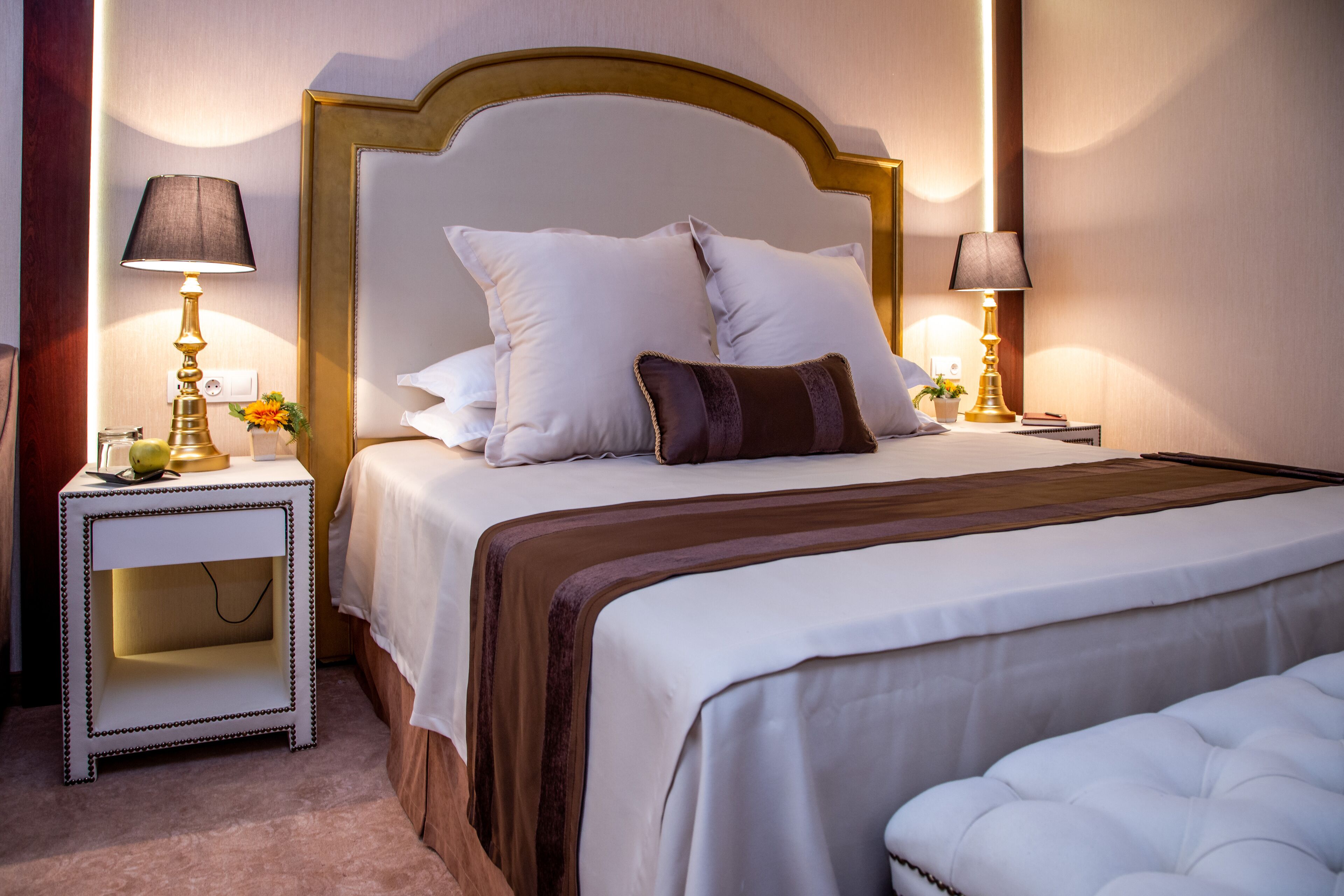 Een netjes opgemaakt bed met wit en bruin beddengoed, twee nachtlampen en een klein nachtkastje met een lamp en een bloemenvaas.