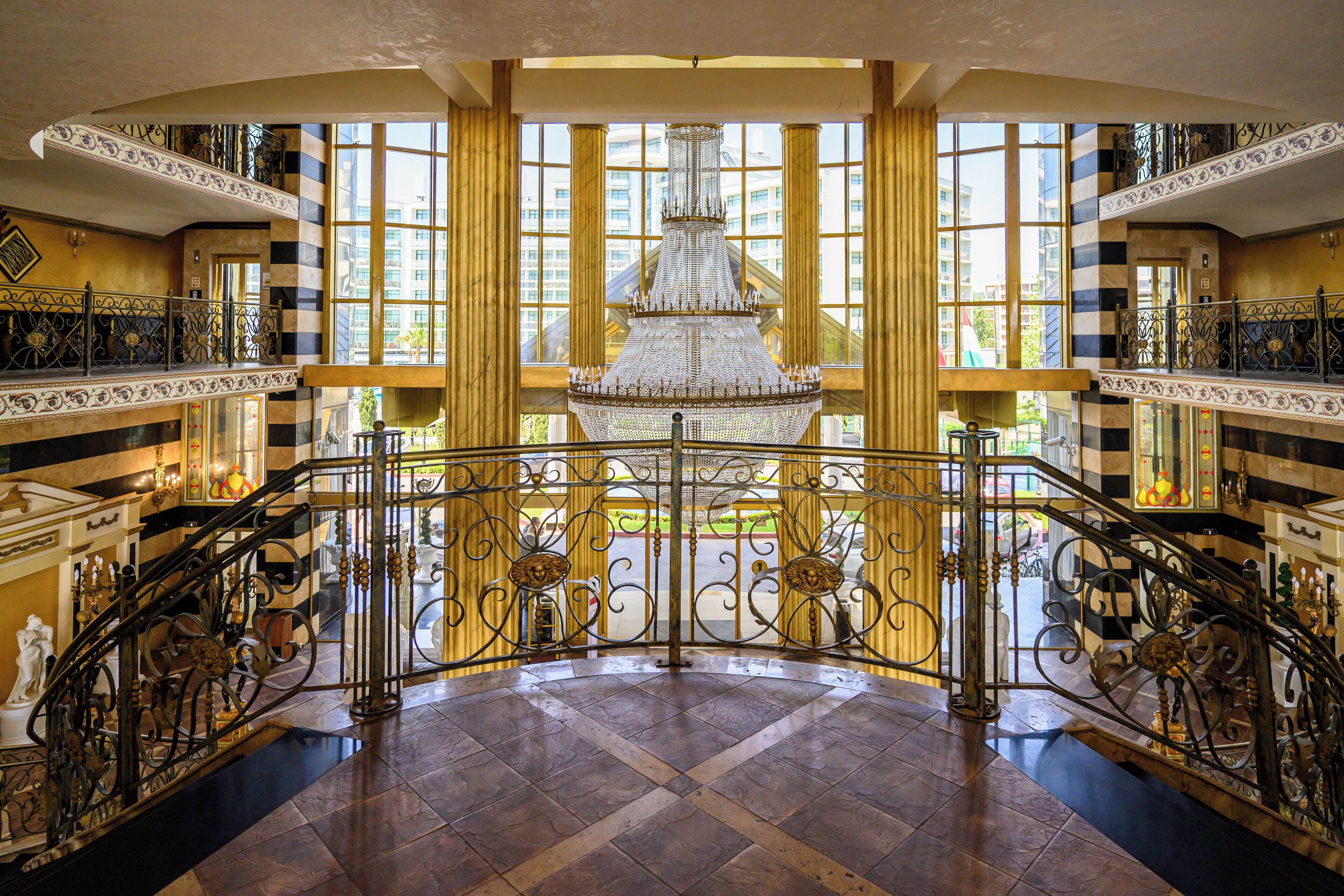 A grand lobby with a large chandelier, ornate iron railings, and a view of the city through tall windows.