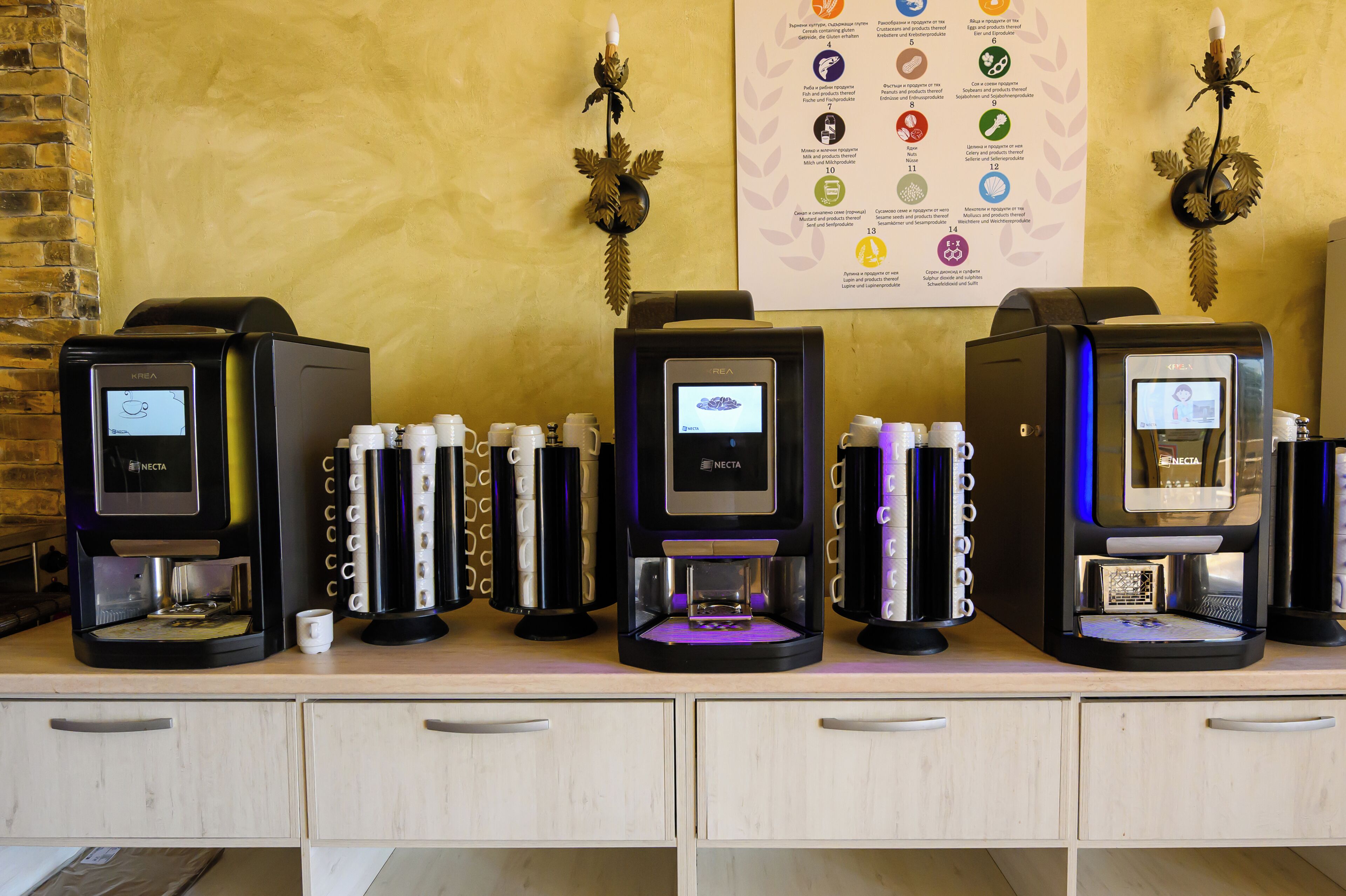 A coffee station with multiple machines and disposable cups, a wall with a food allergy chart, and a brick wall on the left.