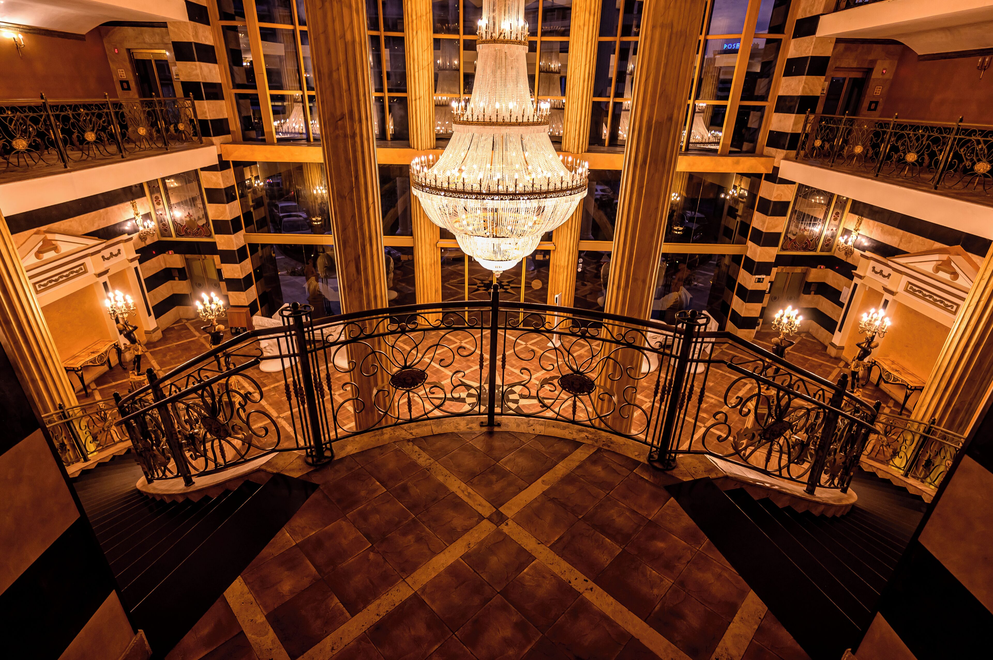 A grand interior with a central chandelier, ornate iron balustrade, and a checkered floor pattern.