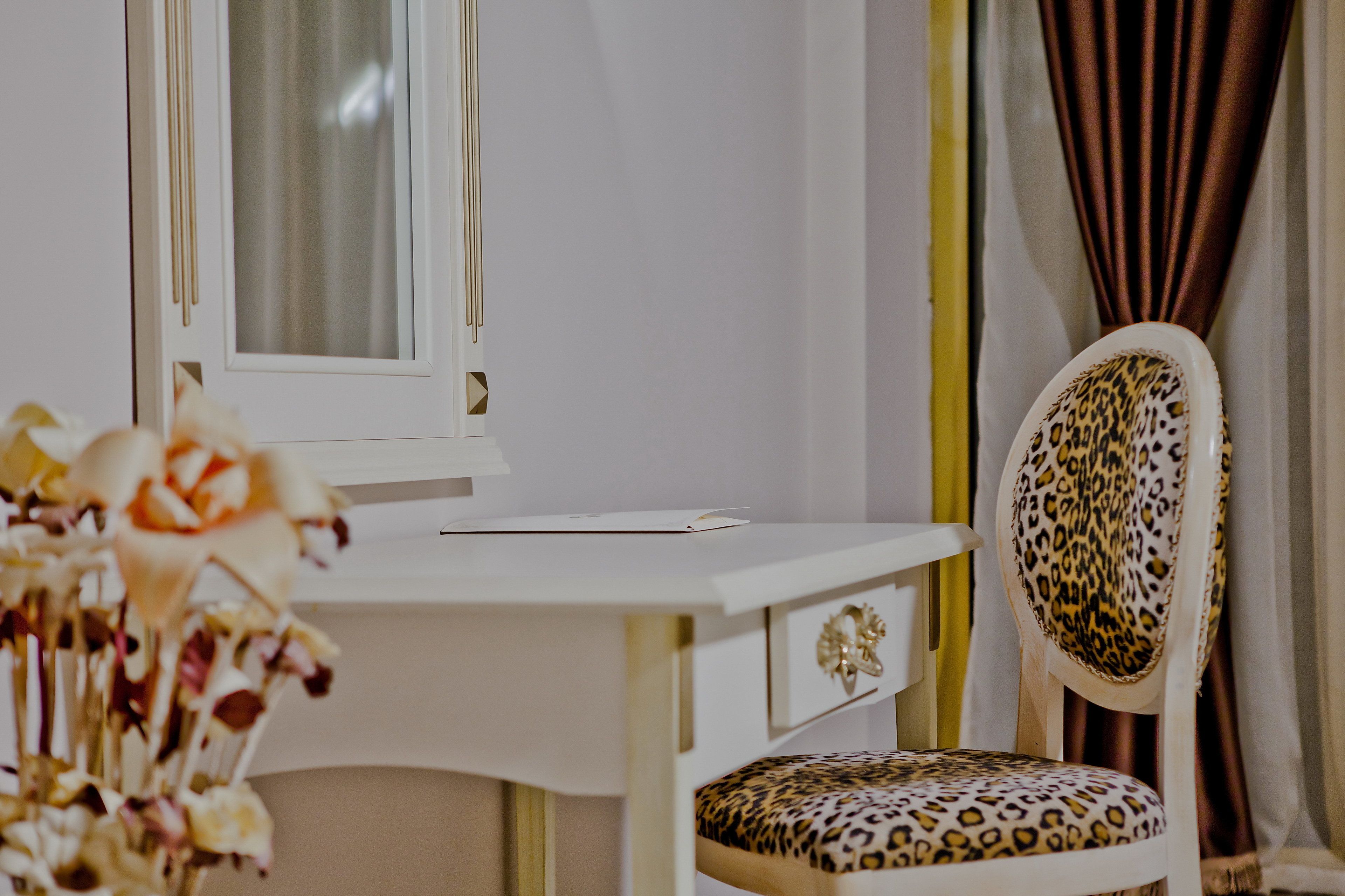 A white desk with a mirror, a chair with a leopard print cushion, and a vase of flowers.