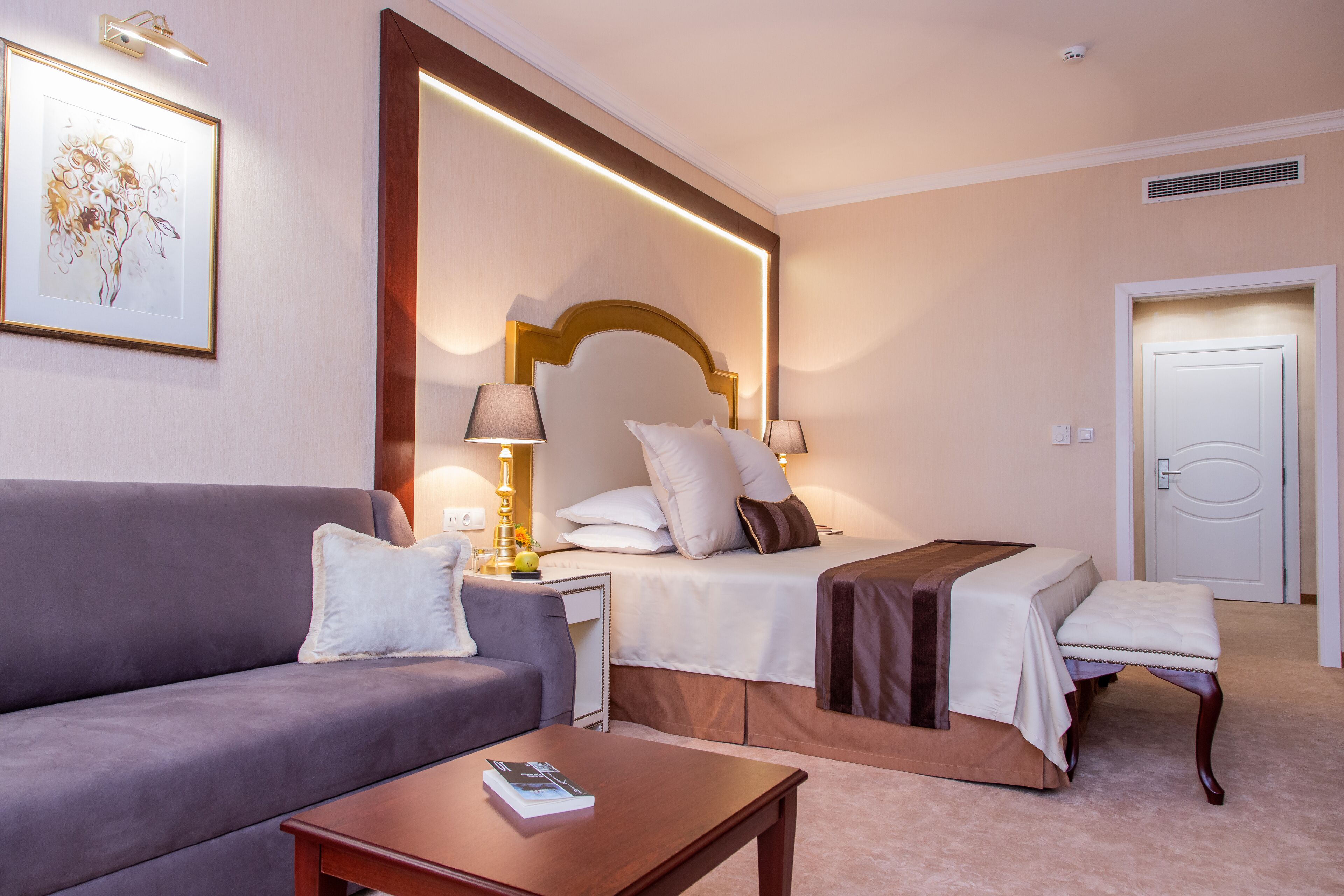 A hotel room with a sofa, a bed with a headboard, a bedside table, and a small table with a book.