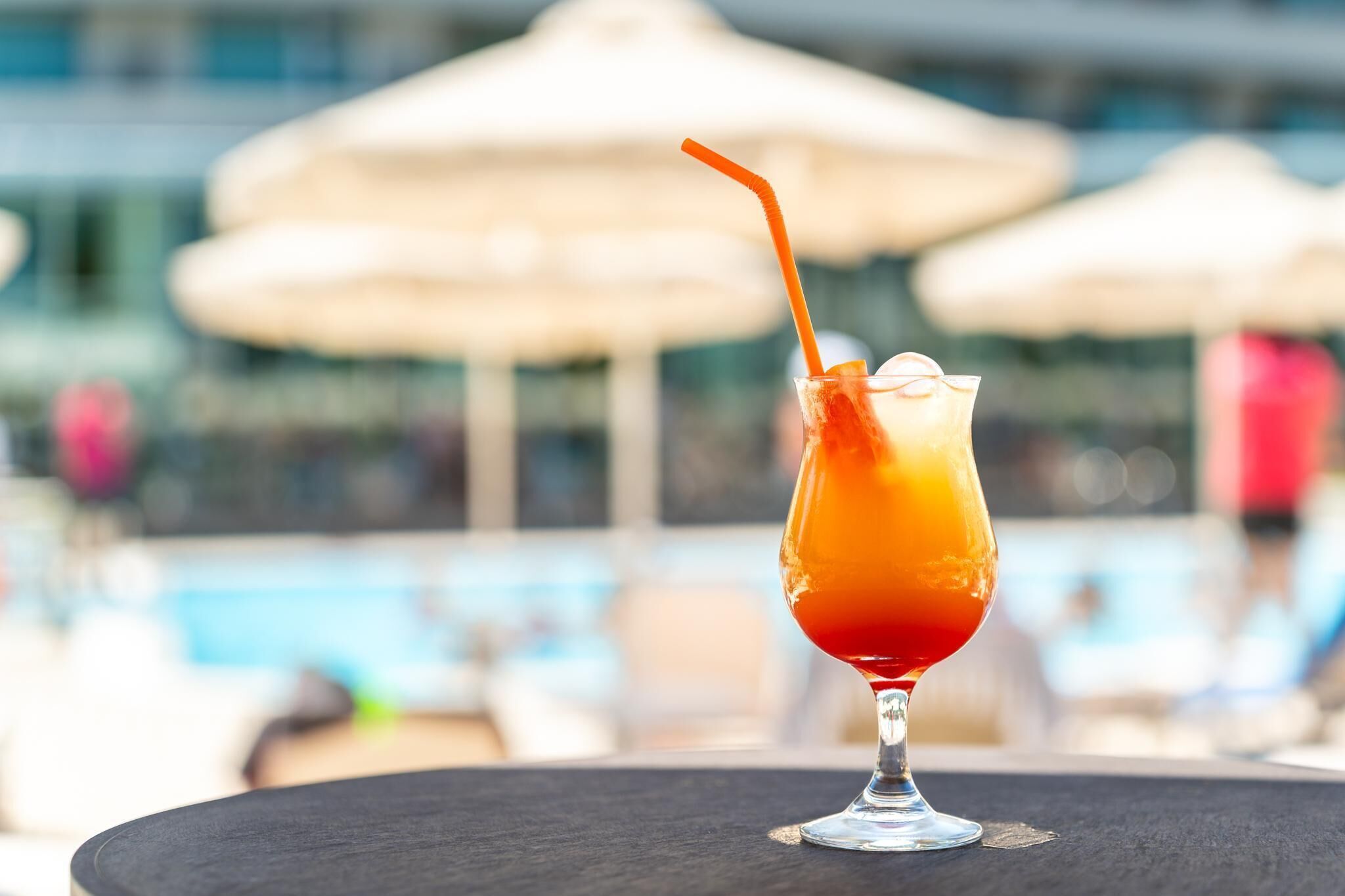 A cocktail in a glass with a straw, placed on a table with a pool and umbrellas in the background.