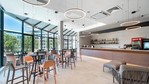 Um café moderno com mesas e cadeiras redondas, área de bar com refrigerador de Coca-Cola e grandes janelas com vista para edifícios e árvores.