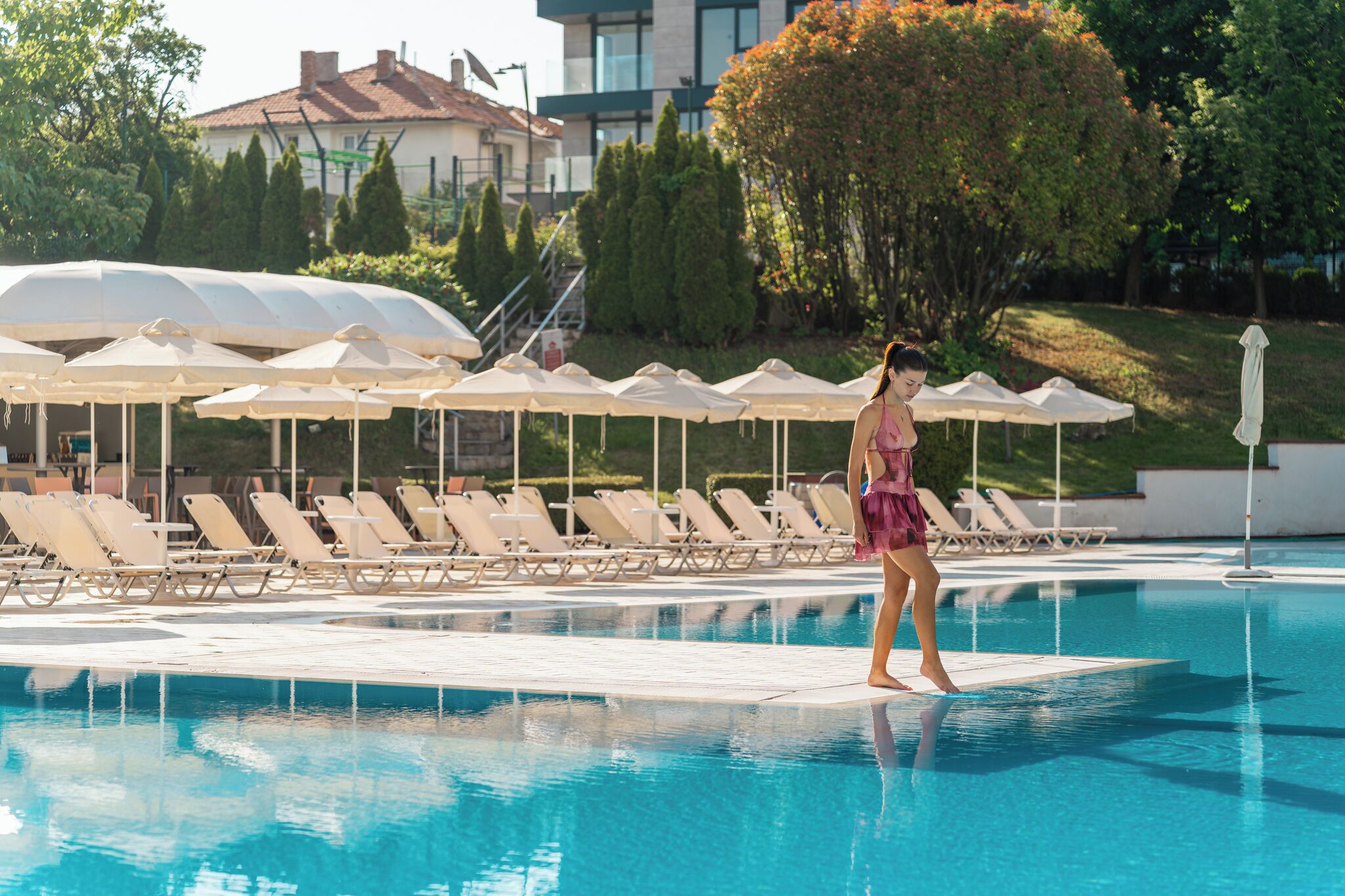 Piscina con sillas de descanso y sombrillas, rodeada de vegetación y edificios.