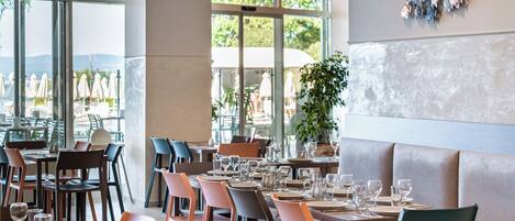 Ein Restaurant mit gedeckten Tischen, Stühlen und einem großen Fenster mit Blick auf Sonnenschirme.