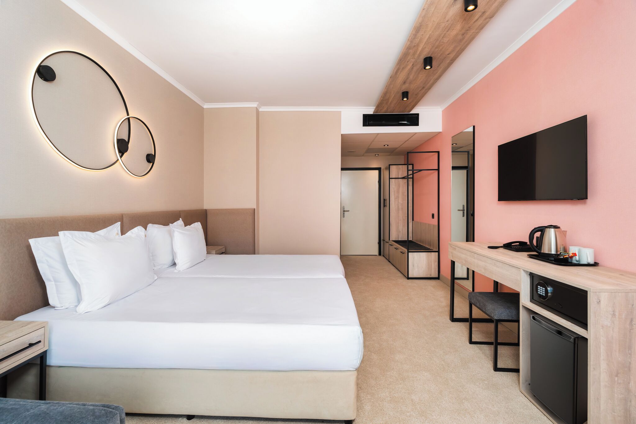 A modern hotel room with a large bed, a desk with a coffee maker, a TV, and a compact kitchen area.