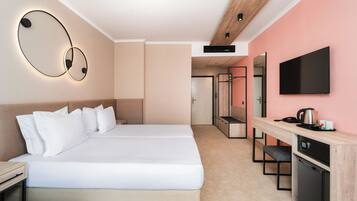 A modern hotel room with a large bed, a desk with a coffee maker, a TV, and a compact kitchen area.