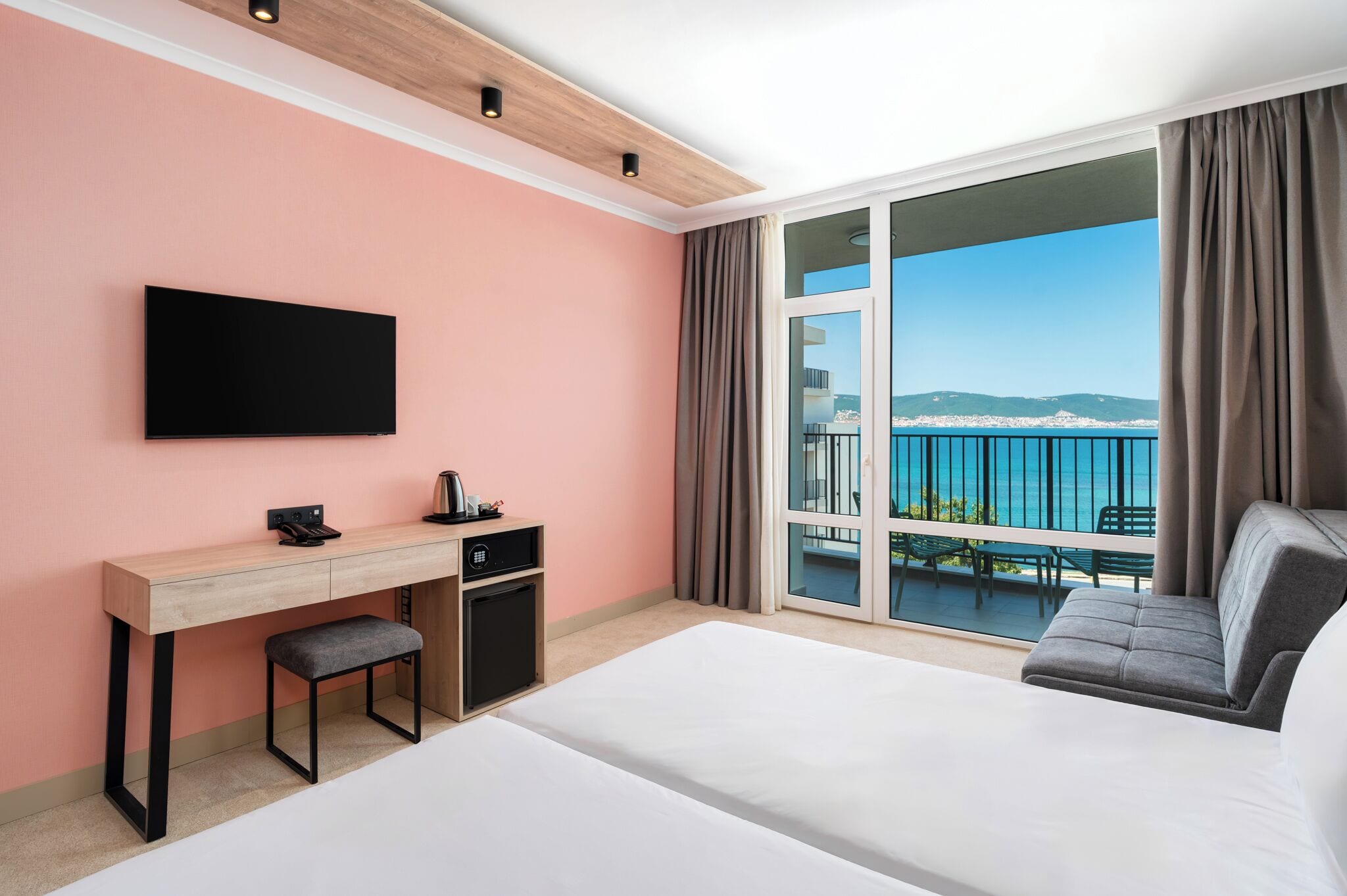 Una camera d'albergo con un letto, una scrivania, una TV, un piccolo frigorifero e un balcone vista mare.