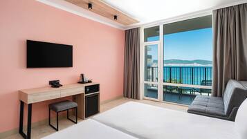 Habitación de hotel con cama, escritorio, televisor, refrigerador pequeño y balcón con vista al mar.