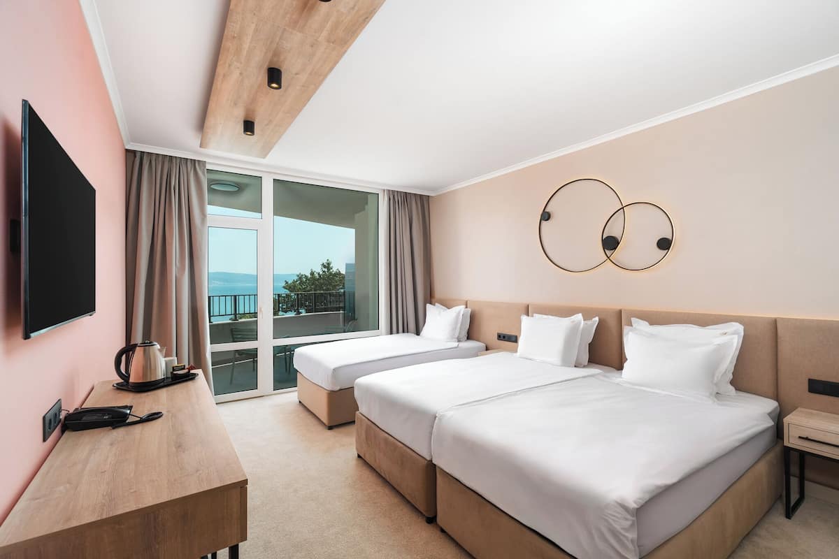 A hotel room with two beds, a desk, a flat-screen TV, and a view of the outdoors.