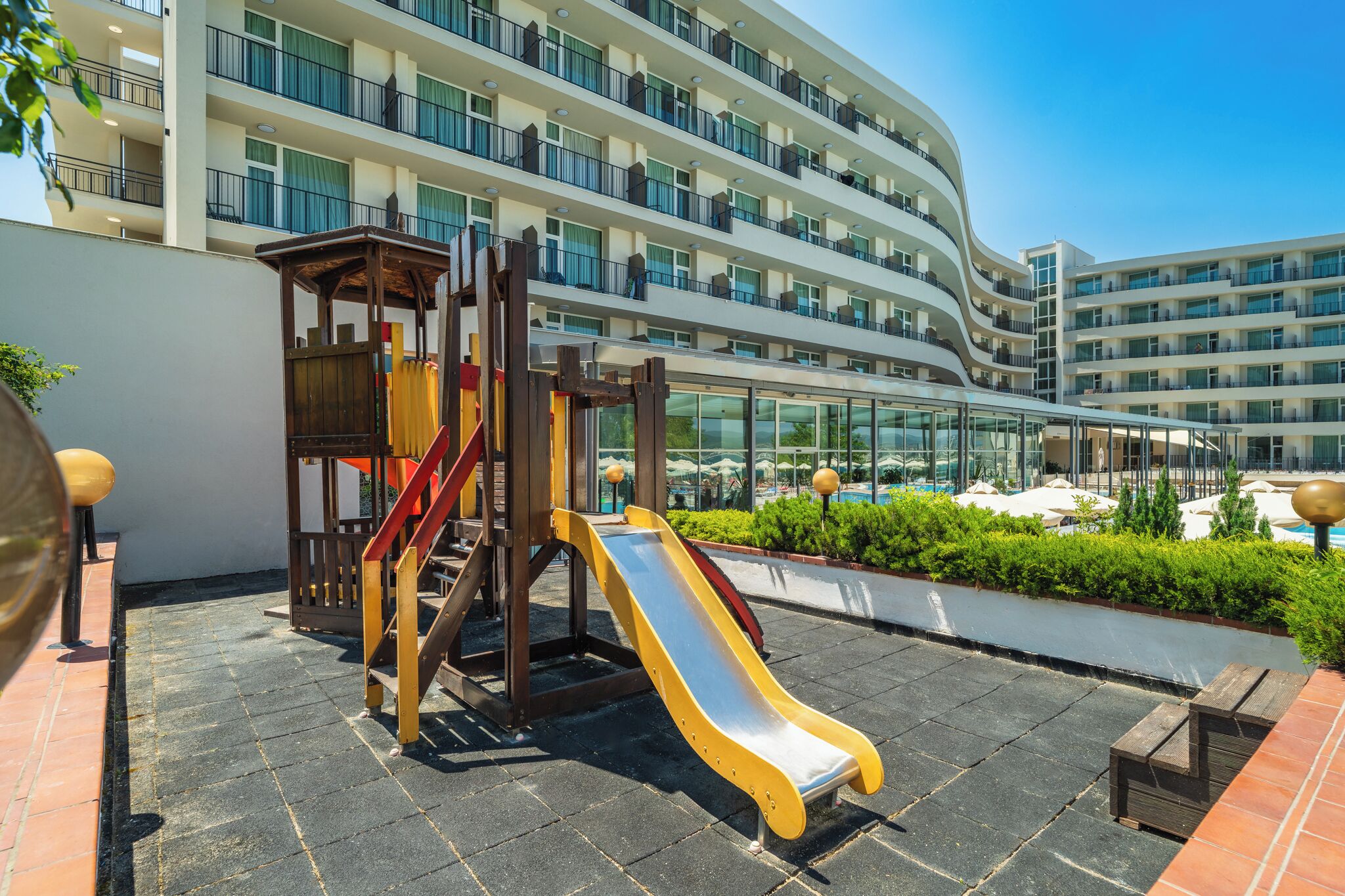 Un parque infantil con tobogán y estructura de escalada, rodeado por una piscina y un edificio de varios pisos con balcones.
