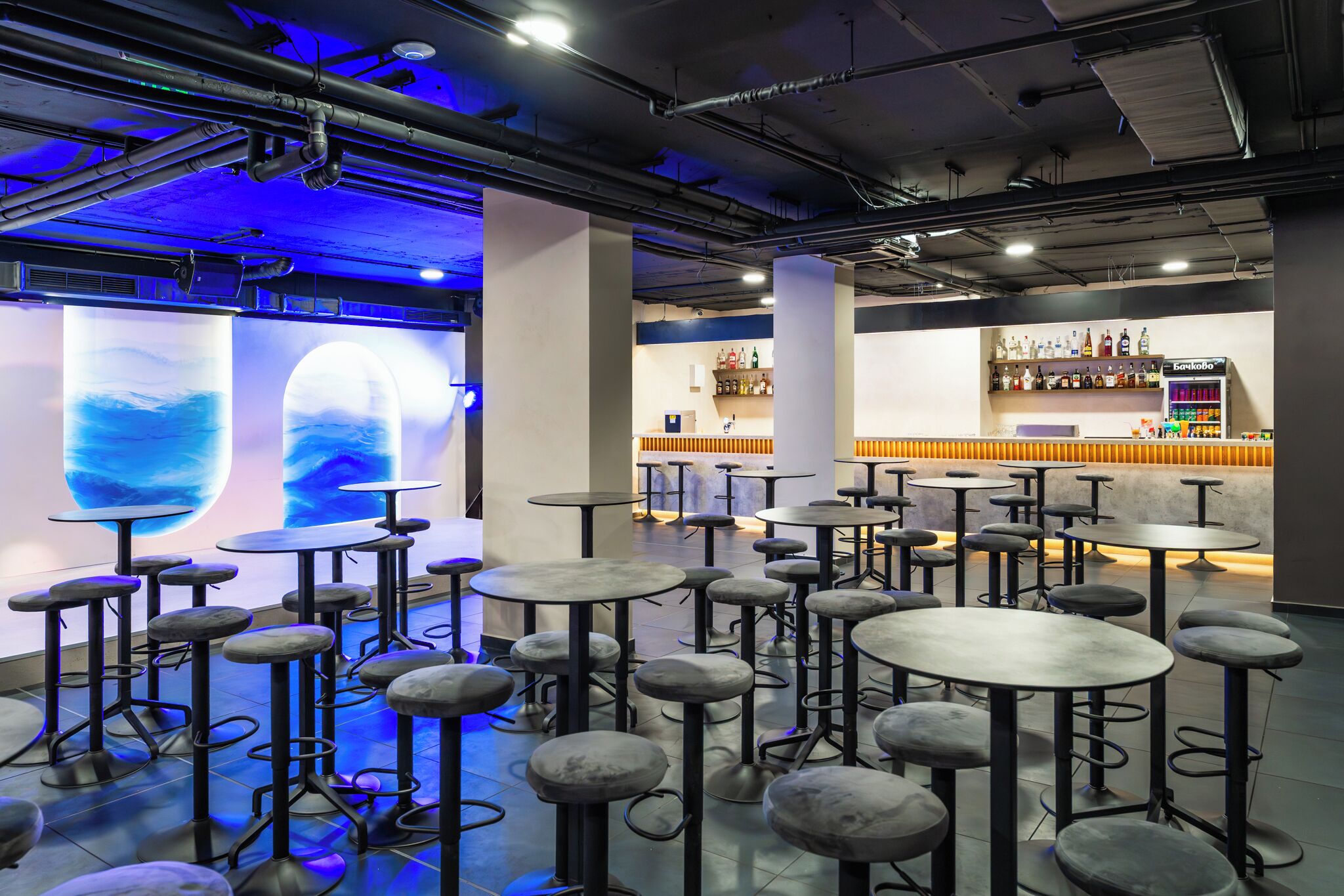 A bar area with multiple stools and tables, a bar counter with bottles, and a wall with a blue light display.