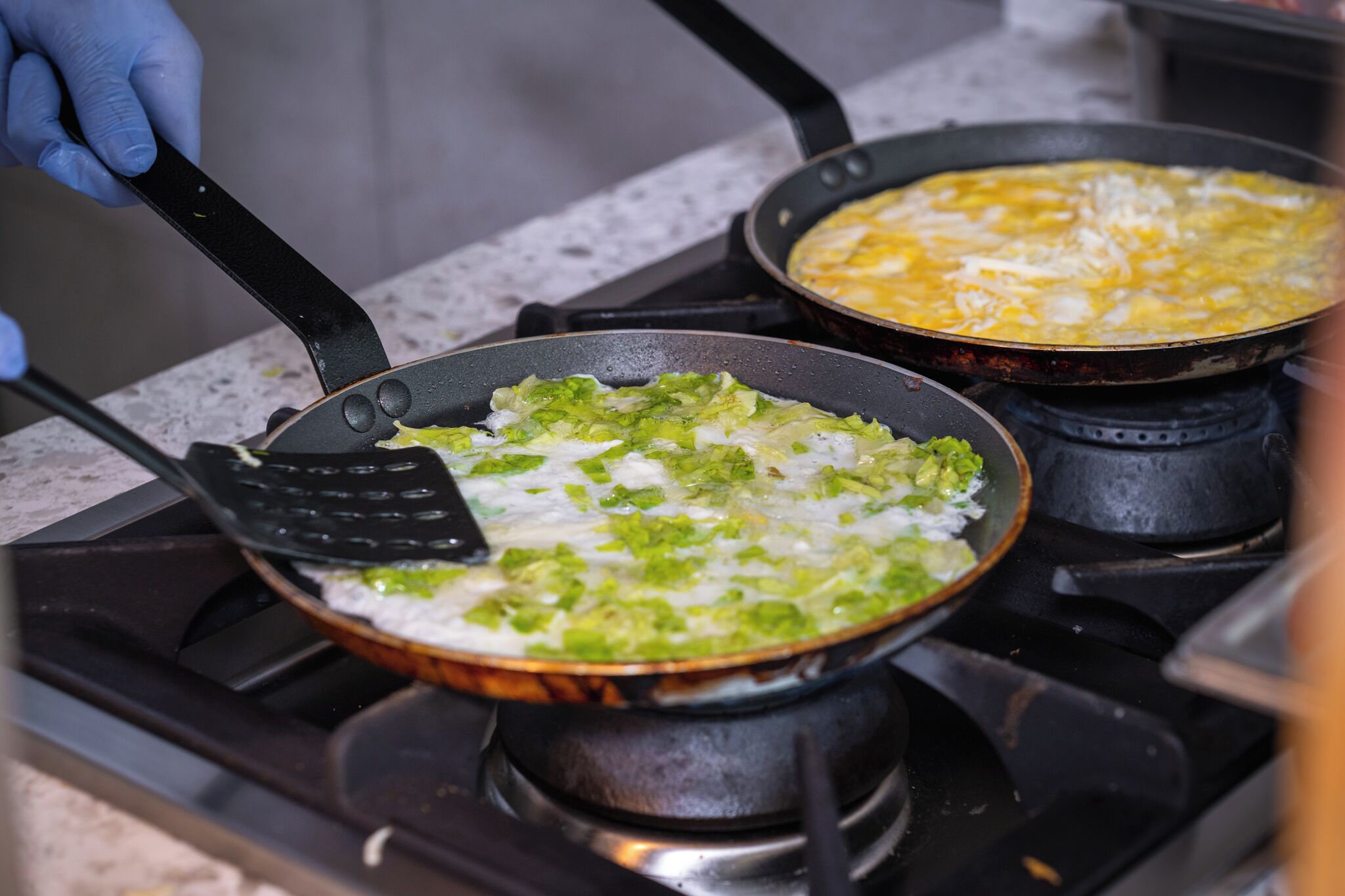 A kitchen with two frying pans on a stove, one containing green vegetables and the other with eggs.