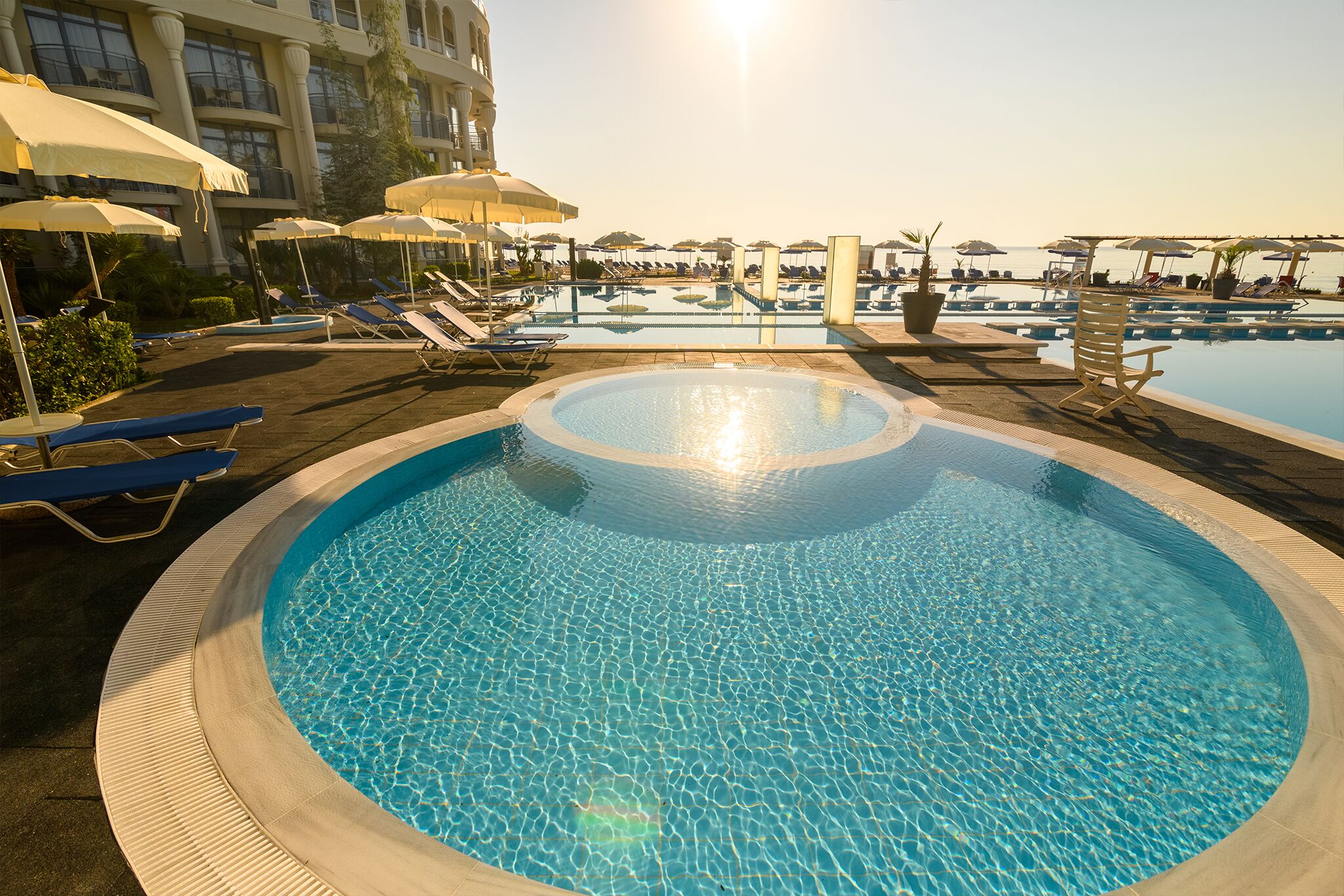 Uma piscina grande e circular com água cristalina, rodeada por espreguiçadeiras e guarda-sóis, com vista para o mar ao fundo.