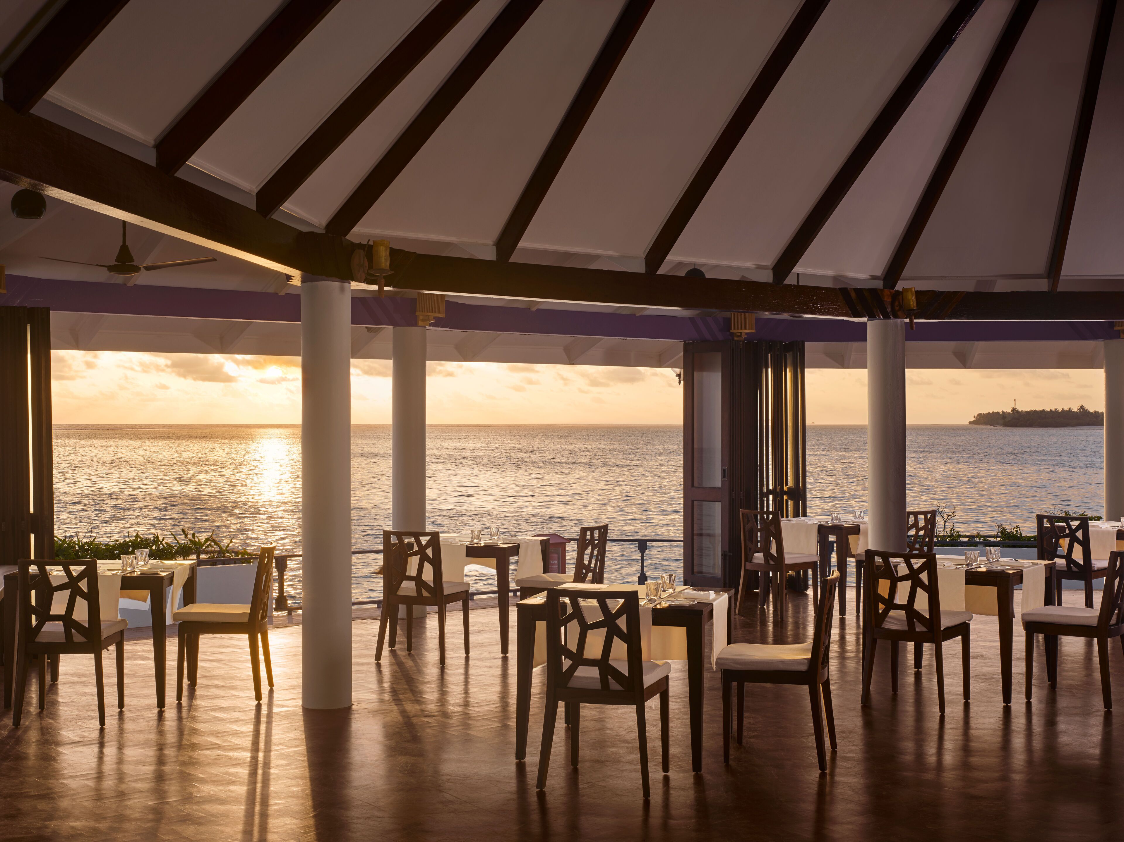 An outdoor dining area with tables and chairs, a ceiling fan, and a view of the sea at sunset.